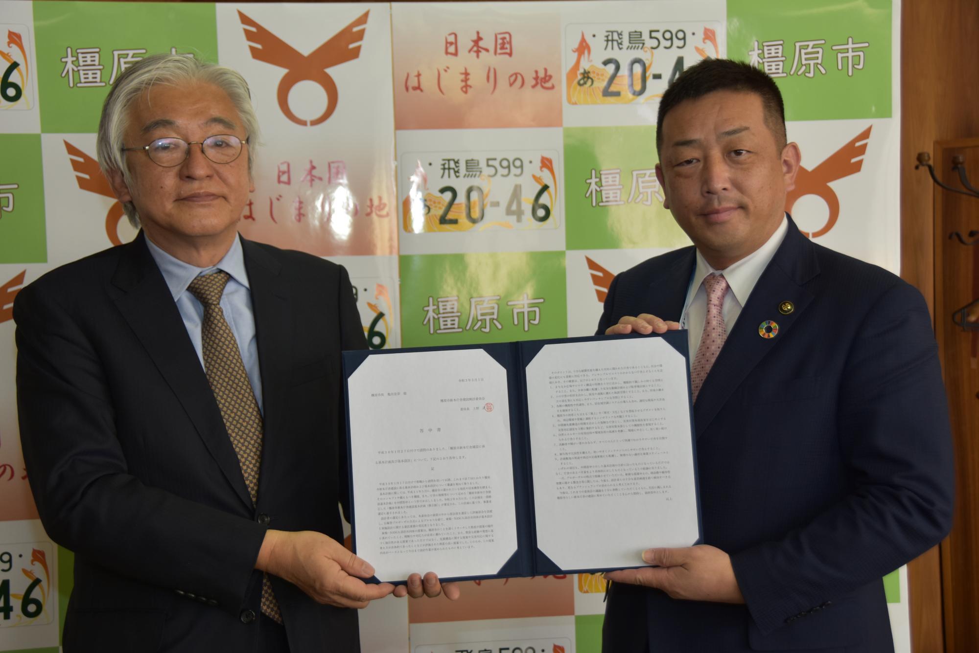 橿原市パネルの前で上野委員長と亀田市長が答申書を持っている様子の写真