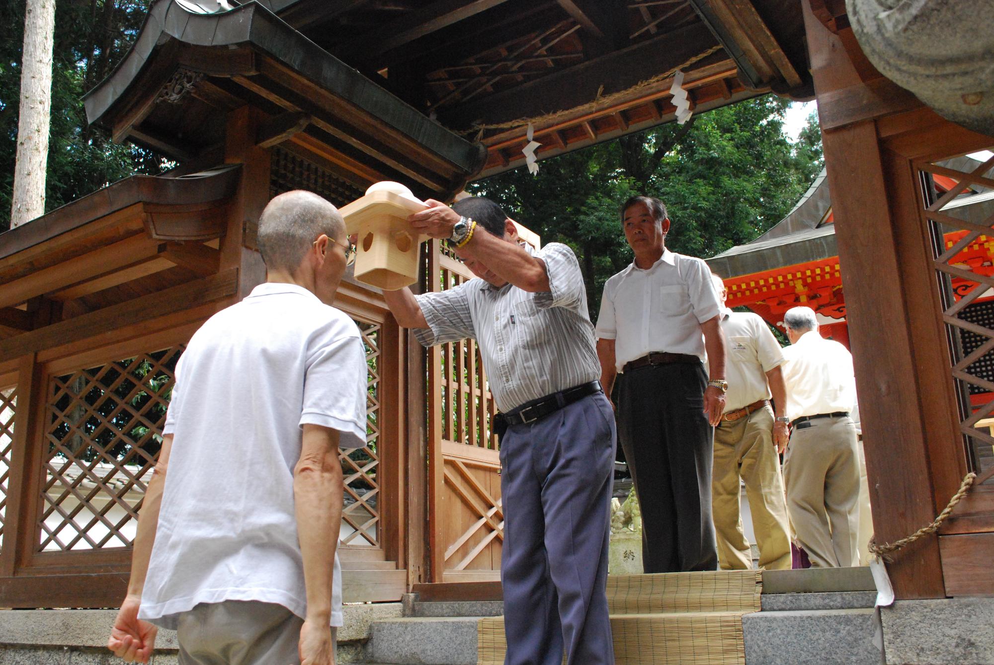 白いシャツを着た男性が門の前で神社に向かって立ち薄いグレーのシャツを着た男性が白いシャツを着た男性に向かって三宝を両手で持って頭を下げている様子の写真