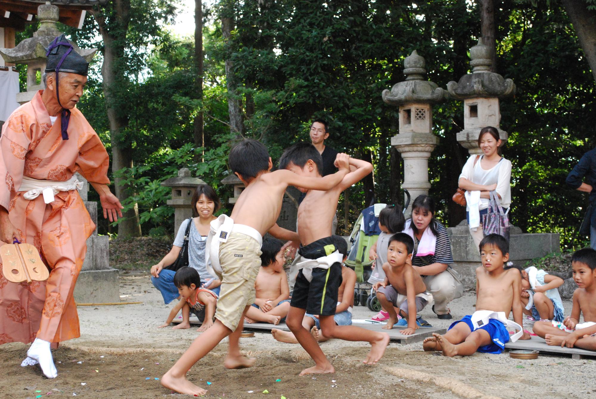 複数の大人や子どもが土俵を囲み、オレンジ色の装束を着た行司が見守る中半ズボンにまわしをしめた二人の子どもが相撲をとっている写真