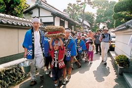 赤いはっぴを着たこどもを先頭に青いはっぴを着たたくさんの子どもたちがわらで作られた大きな蛇を住宅街の中をひいて歩いている様子の写真