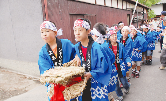 青いはっぴを着て頭にはちまきを巻いたたくさんの子どもたちがわらで作られた大きな蛇をひいて町なかを歩いている様子の写真