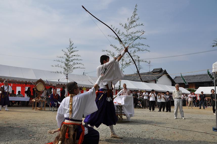 本薬師寺跡で白いタープテントが並び広場の中央で男性が大きな弓を引いている様子とそれをたくさんの人たちが見物している写真