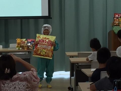 工場のユニフォームを着た人が大きなポテトチップスの袋をもって講義を受ける人たちの前に立っている写真