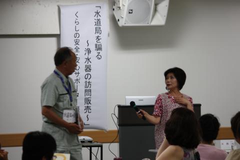 「水道局を騙る~浄水器の訪問販売~」の垂れ幕の前で講演者2人が話している写真