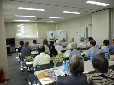 一番前で話をする講師と机が並べられた室内で話を聞く多くの人の写真