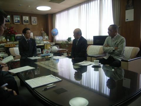 テーブルを囲んで亀田市長から一次答申を受ける川上会長と米田副会長の写真