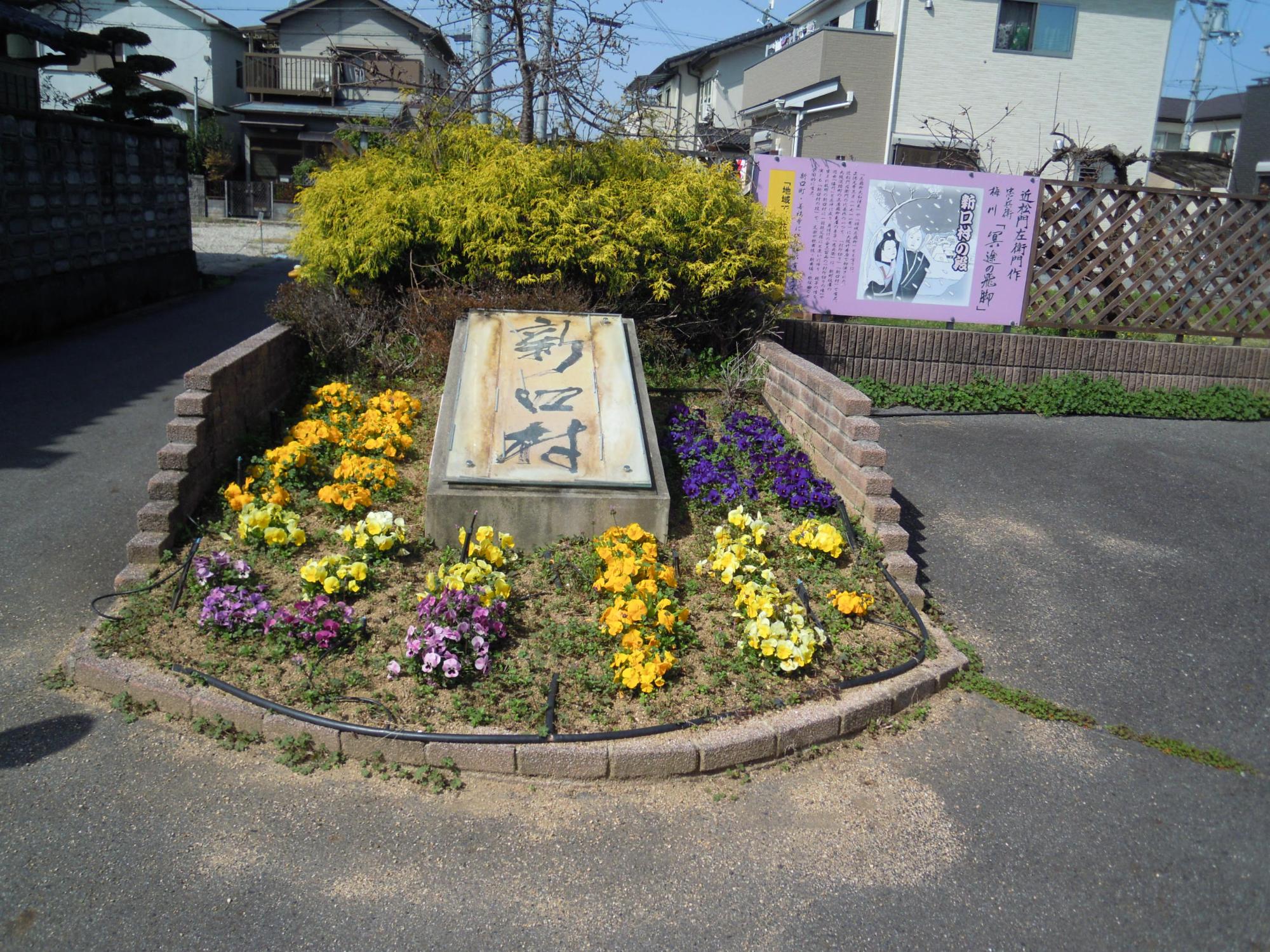 新口村と書かれている石碑と黄色と紫色の花が咲いている忠兵衛碑花壇の写真