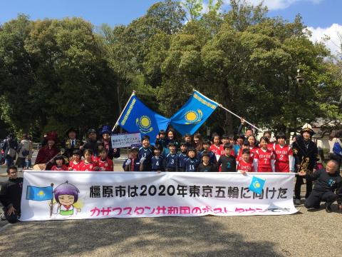 「橿原市は2020年東京五輪に向けたカザフスタン共和国のホストタウンです」と書かれた大きな横断幕やカザフスタン国旗をもった、複数のパレード隊の人たちの集合写真