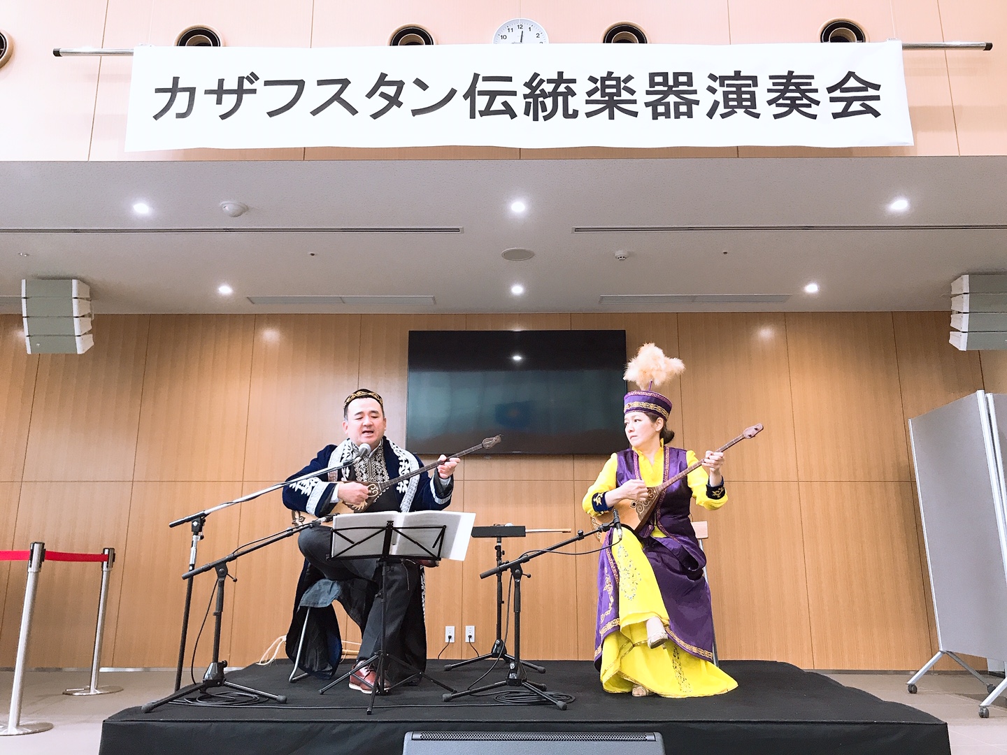カザフスタン伝統楽器演奏会と書かれた垂れ幕の下で、伝統楽器を演奏しながら歌っているイナーラ・セリクパエヴァさんと高橋直己さんの写真