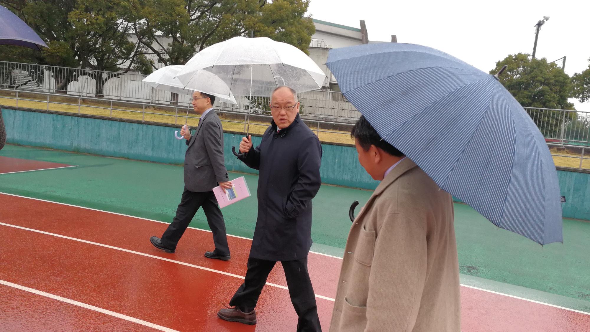 笠井さんと男性二人が傘をさしてスポーツ施設の外を歩いている写真