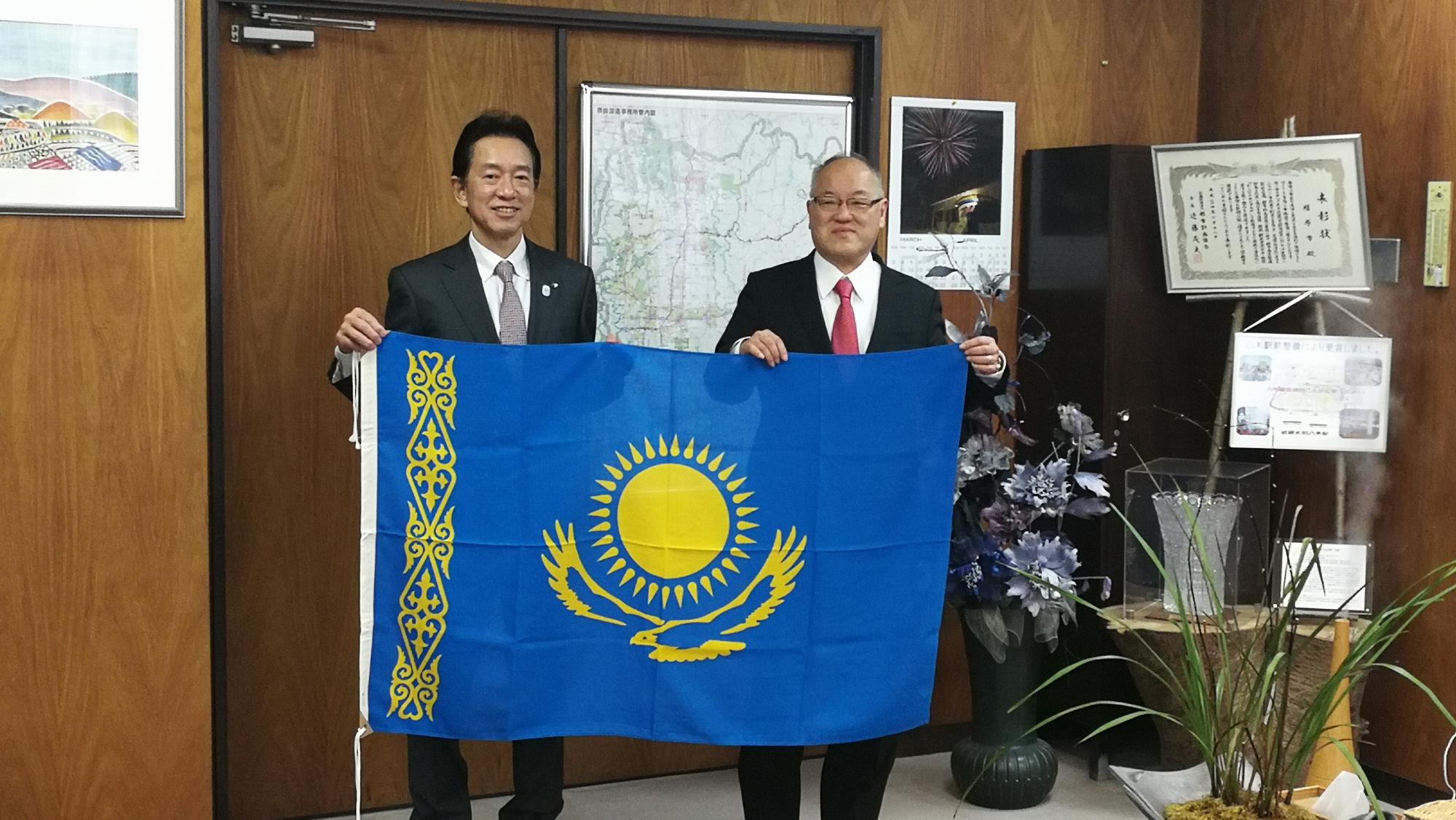 森下市長と笠井さんがカザフスタンの国旗を挟んで両端に立っている写真