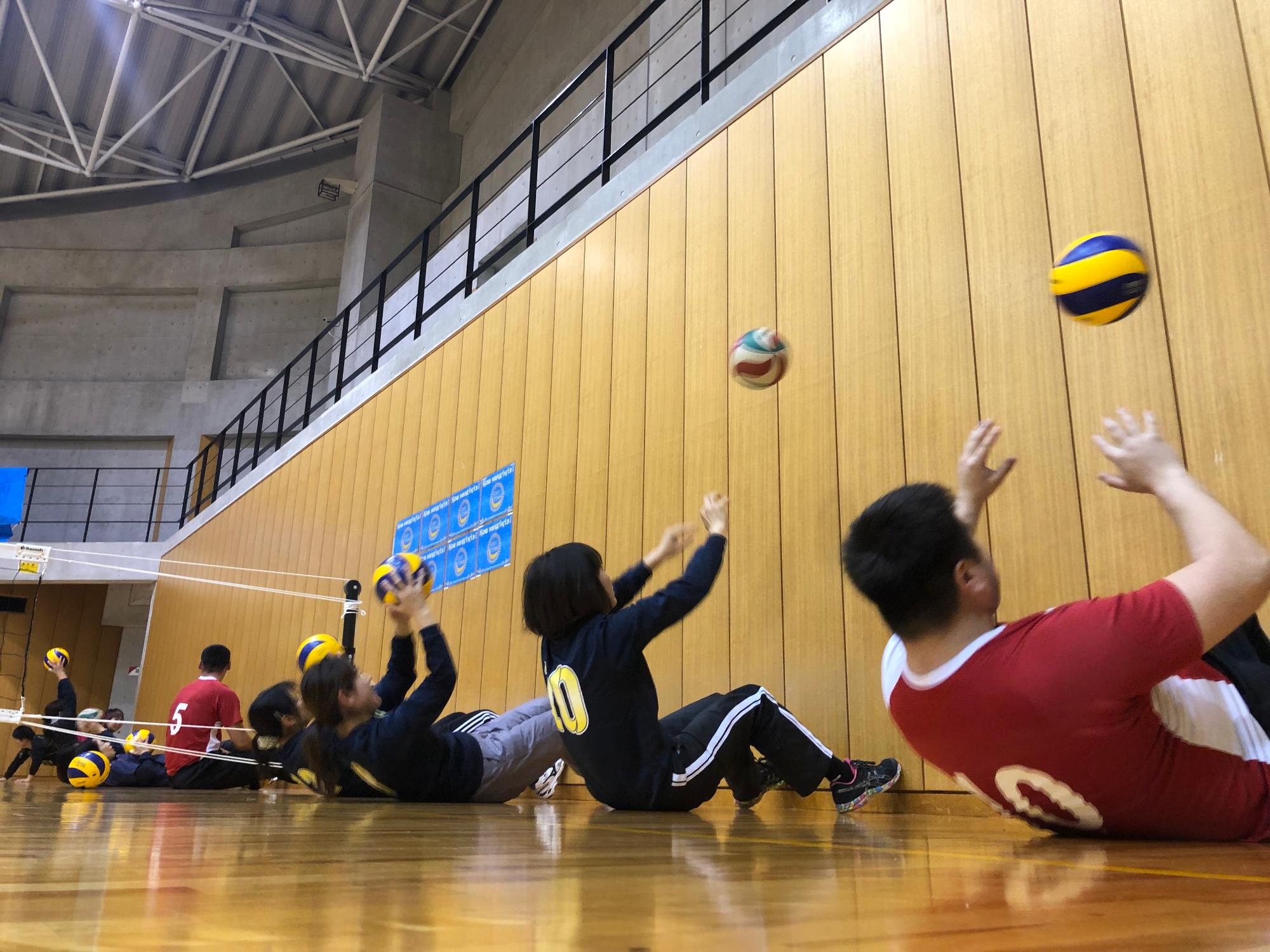 体育館の壁沿いに座って真上にボールを投げる参加者の写真