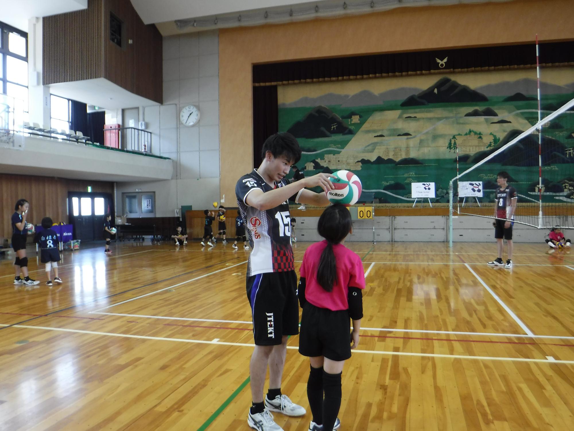 選手が生徒に体育館でバレーボールを持ってレクチャーしている写真