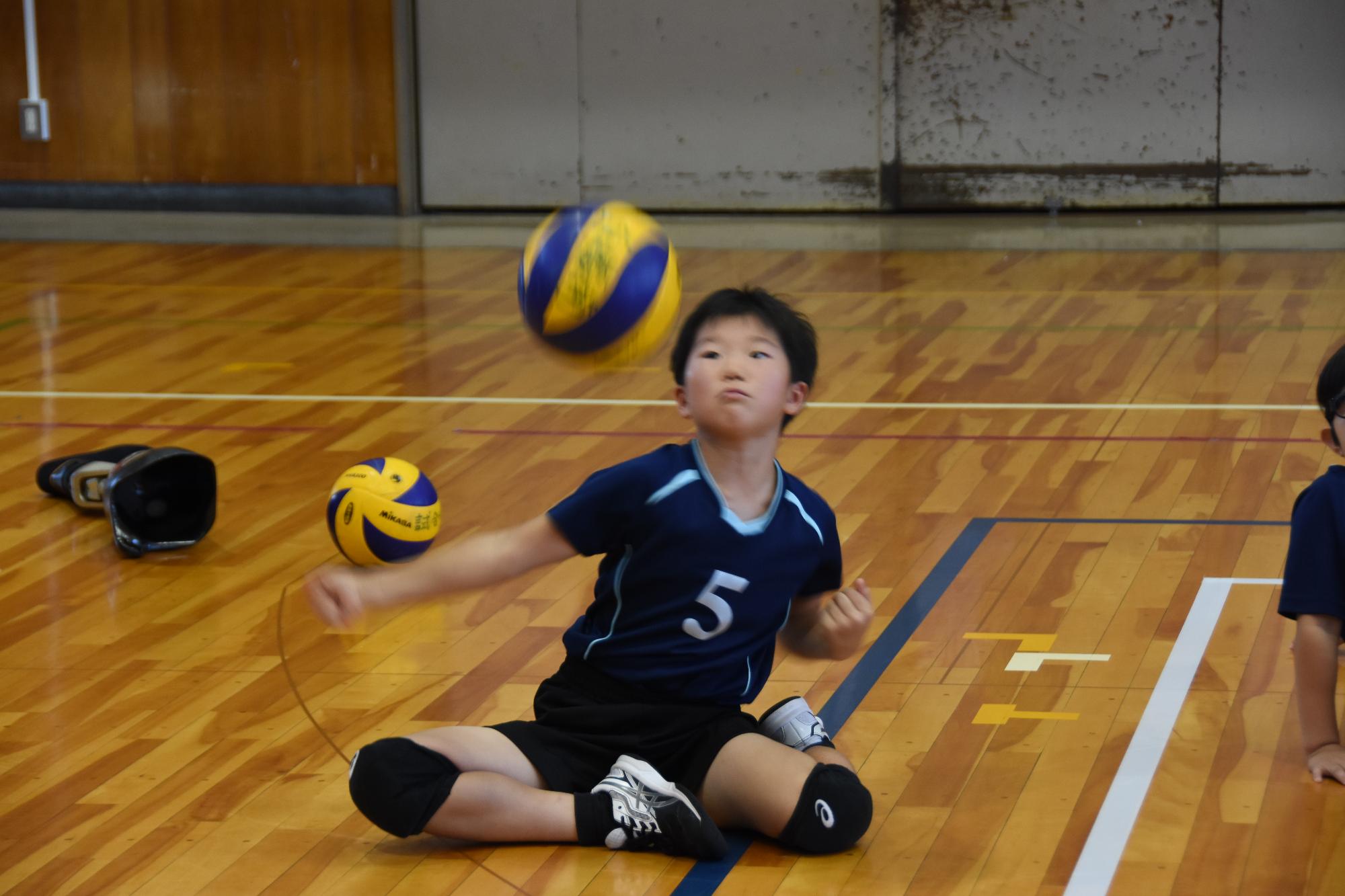 男子生徒が体育館でシッティングバレーボールに挑戦している写真