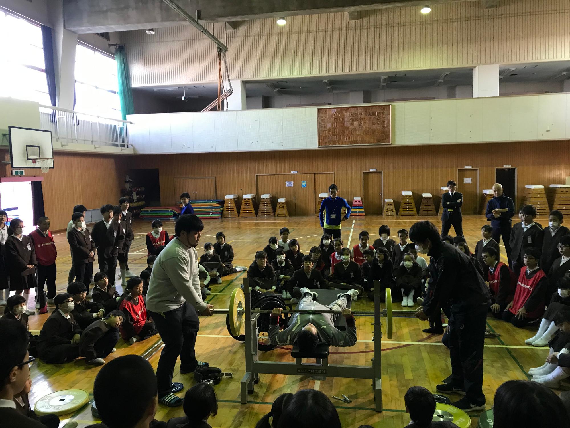 車椅子の男性選手が台の上でパワーリフティングをしている様子を多くの児童が見ている写真
