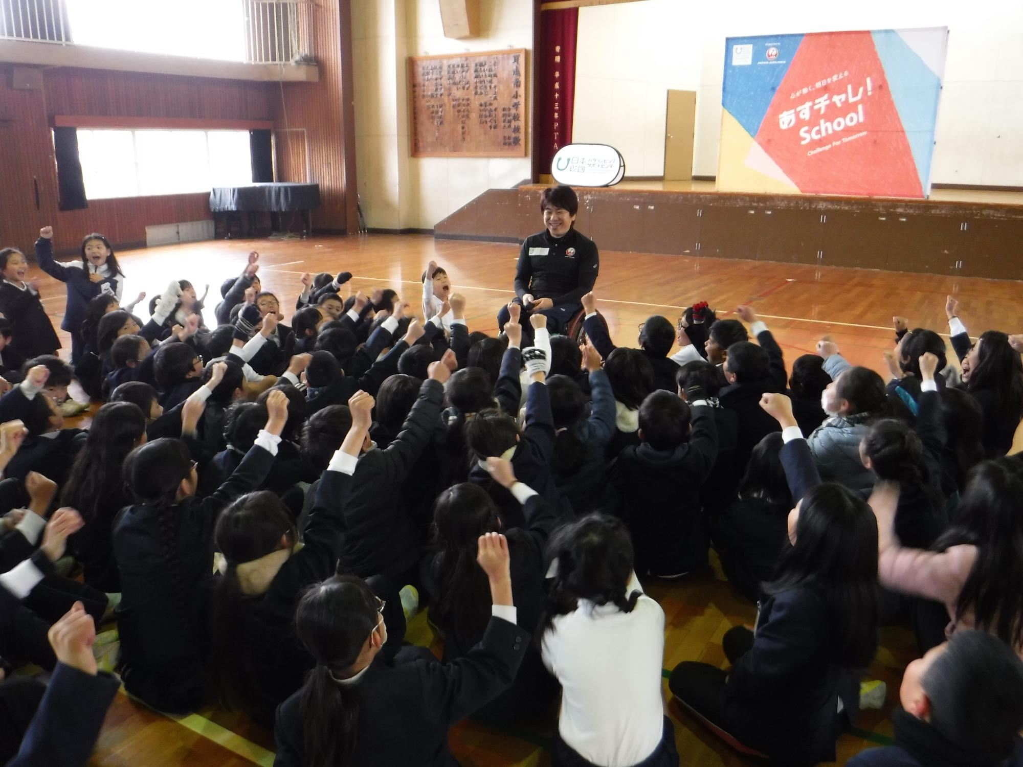 パラリンピックの車いすバスケットボール日本代表チームの元キャプテン、根木慎志さんを前に大勢の小学生たちがガッツポーズをしている写真