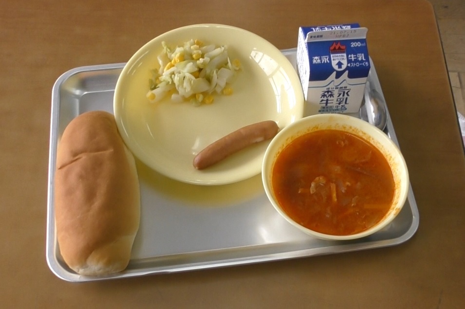 机に乗った銀のお盆の上にボルシチ、紙パックの牛乳、パン、ウインナーとサラダが乗っている写真。