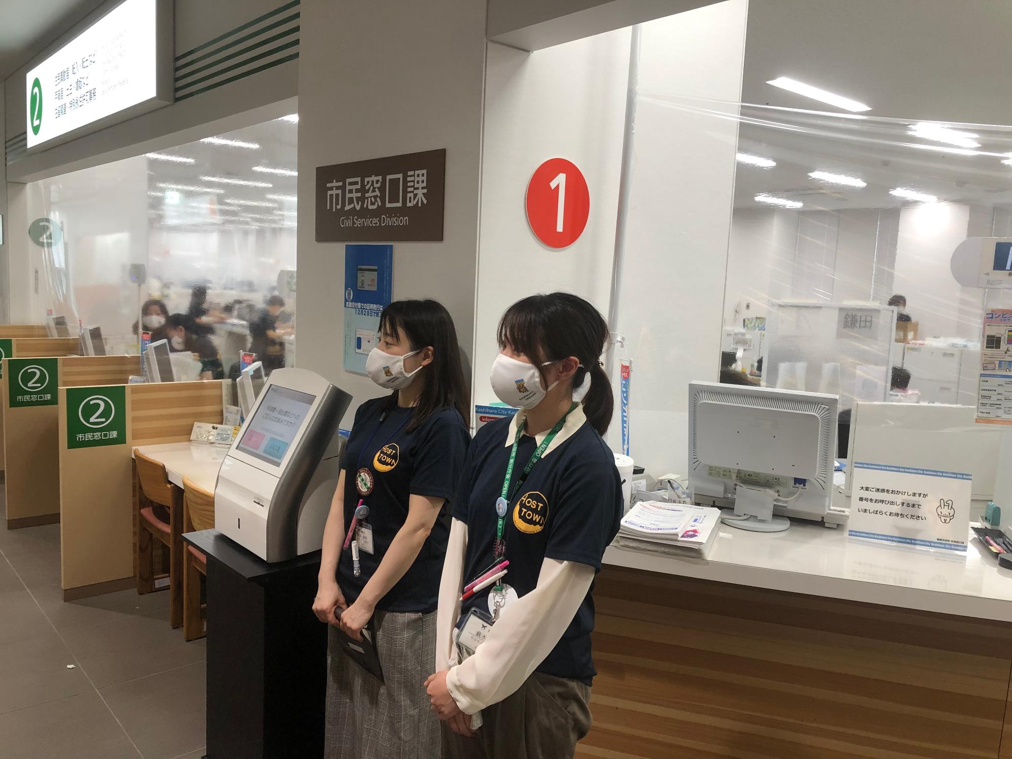職員の女性2人が、橿原市役所市民窓口課の受付の前でホストタウンPRマスクとTシャツを着用し立っている写真。
