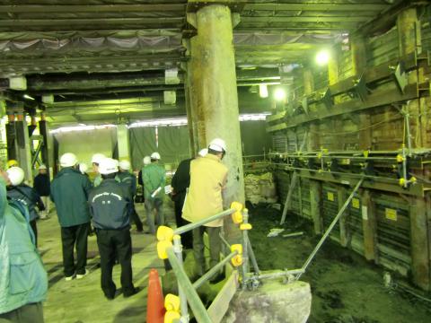 工事現場屋内で説明を受けるヘルメットをかぶった議員達の写真