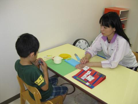 おままごとセットを前にその道具を両手で持っている少年と向かい合って支援をしている女性の支援員の写真