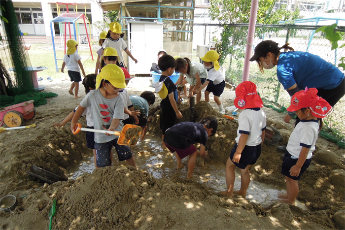 園庭の砂場で園児が穴を掘って温泉作りをして遊んでいる写真