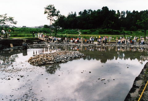 真ん中に石をたくさん盛ってつくられた島がある池の周りにたくさんの人が集まって見ている様子の写真
