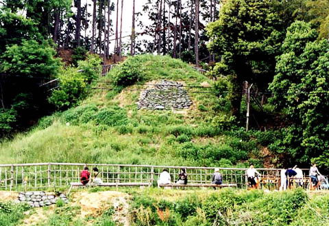 小高い山の斜面の草や木に囲まれたところに四角く石が並べられたキトラ古墳とそれを眺めるように置かれたベンチに座っている人々の写真