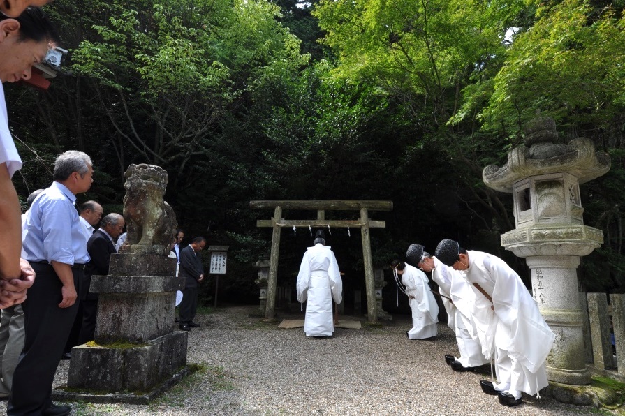 鳥居の前に白い袴を着た1人の神官が立ちその横で3人の神官がお辞儀をして複数のスーツを着た男性が頭を下げている様子の写真