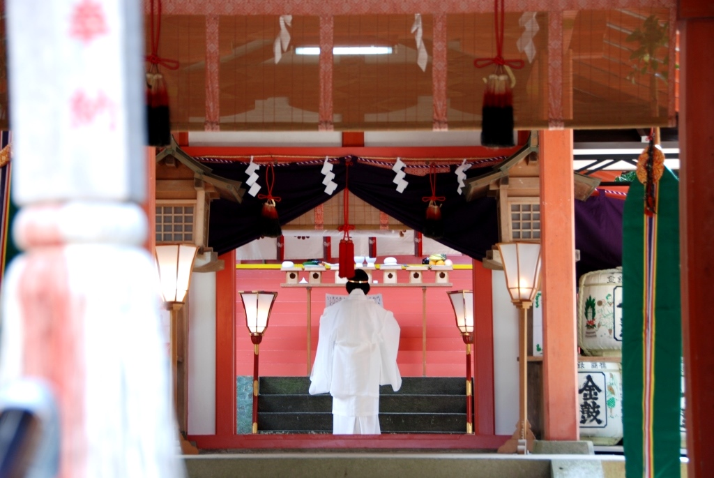 白い袴姿の人が朱色の神殿の奥にある神棚の前に立ち祝詞をとなえている様子の写真