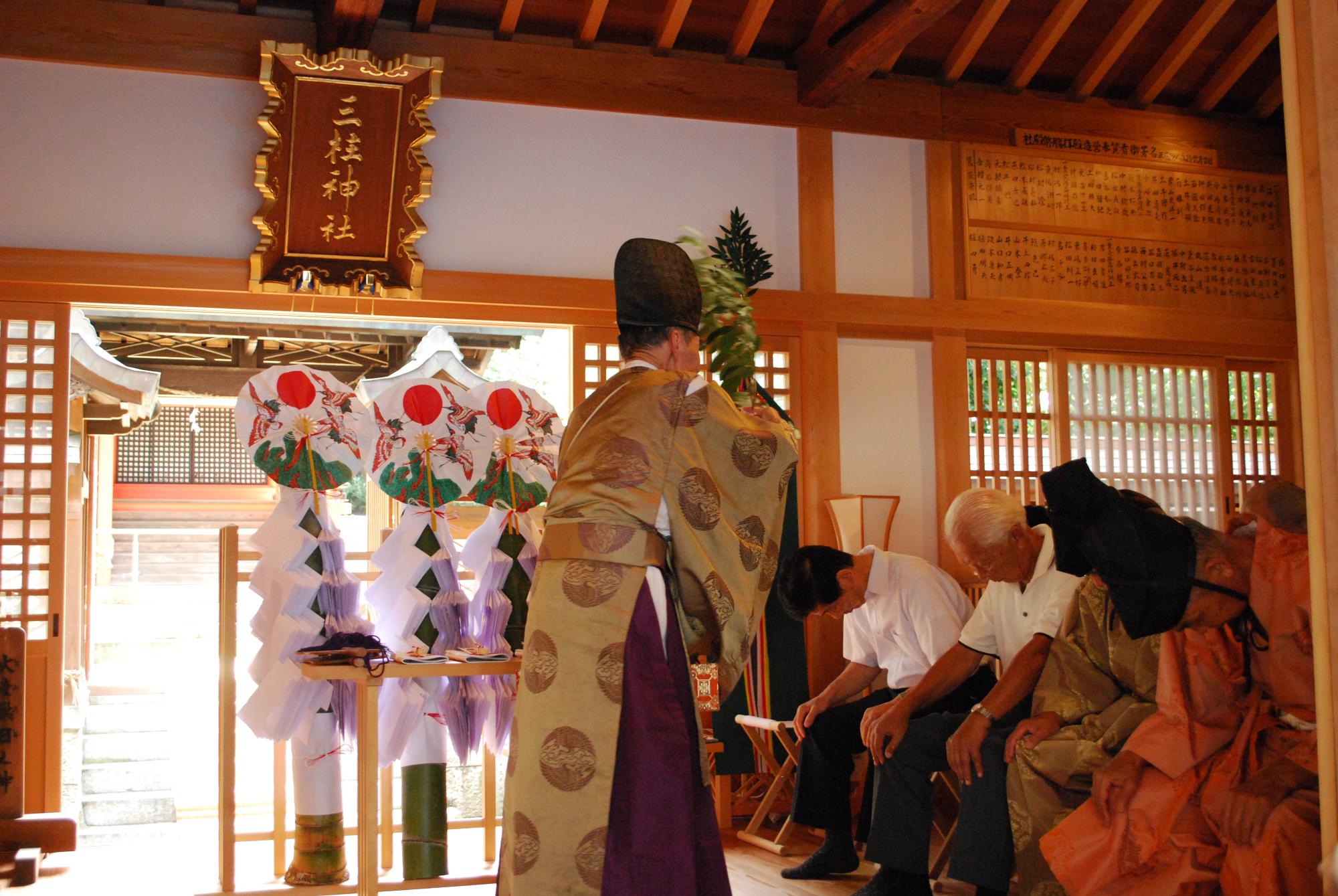 神社の中でオレンジ色の装束を着た行司、うすい茶色の装束を着た行司、白い半袖シャツ姿の男性が祈祷を受けている様子の写真