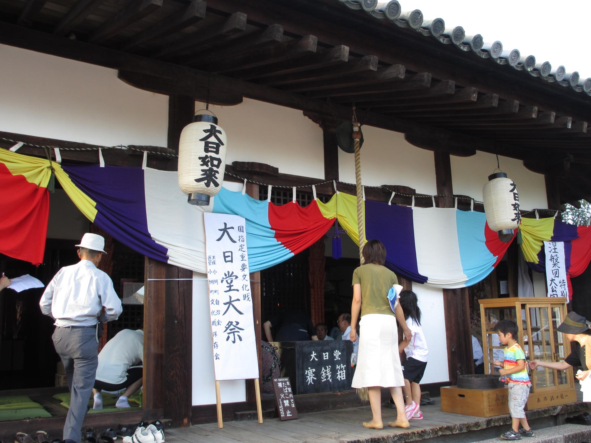 寺の入り口に立てかけられた「大日堂大祭」と書かれた看板の横にある賽銭箱の前で大人の女性と子どもがお参りをしようとしている様子の写真