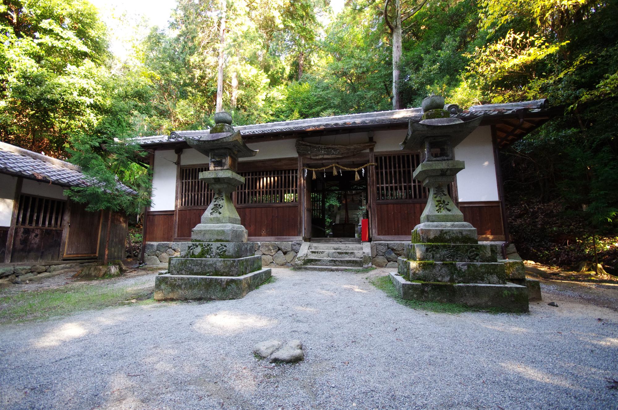 林の中に建っている小さな神社の前に、背の高い石の灯籠が2つ置かれている写真