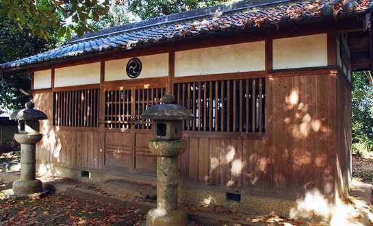 木の壁と扉に瓦屋根の小さな建物と、石の灯籠が2つ入り口に飾られている写真