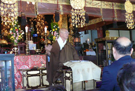 お堂の中にある祭壇の前で茶色の法衣を着た男性が話をしている様子の写真