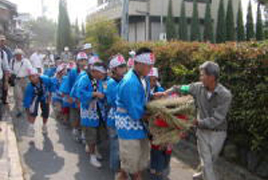 青いはっぴを着たたくさんの子どもたちと先導している男性がわらで作られた大きな蛇の頭部を抱えて歩いている様子の写真