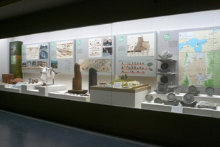 壁のガラス展示の中に、大小様々な土器と、背後の壁に地図や解説のプリントが貼られている写真