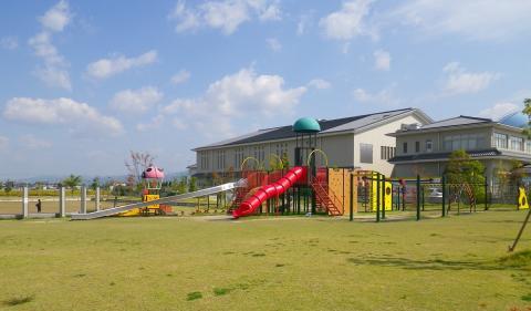 広々とした芝生とともに奥に複合施設と手前に遊具が写っている晴れた日の東竹田近隣公園の写真