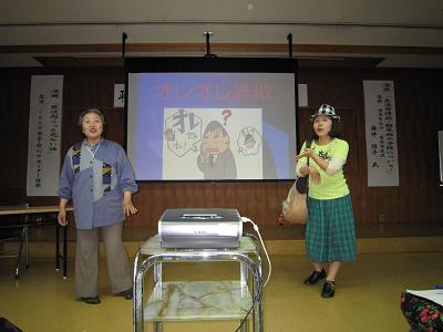 嫁役の女性が左手を姑役の女性の方に向けている写真