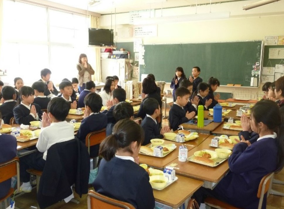 向かい合うように机を突き合わせて座る生徒たちが給食を前に「いただきます」と合掌している様子