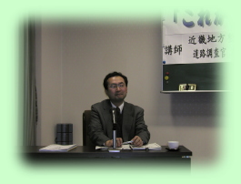 机の前で座っている板倉信一郎氏の写真
