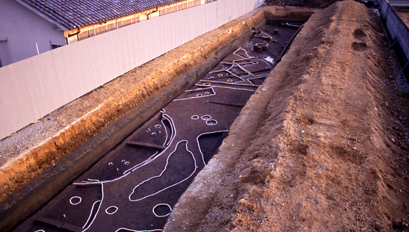 「下明寺竪穴住居」跡の写真。長方形に掘られた地面に丸や四角などの白い枠が示されている