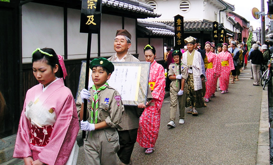 桃色の着物や茶人の服装など和服の男女が行進している今井町並み散歩の様子を撮影した写真