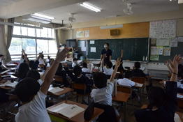 低学年の児童たちが手を挙げて先生の質問に答えようとしている写真